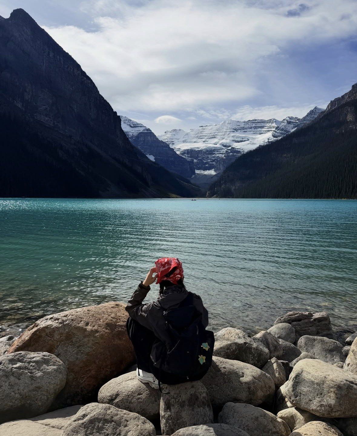 Hiker with backpack sitting on shoreline boulders facing Lake Louise glacier