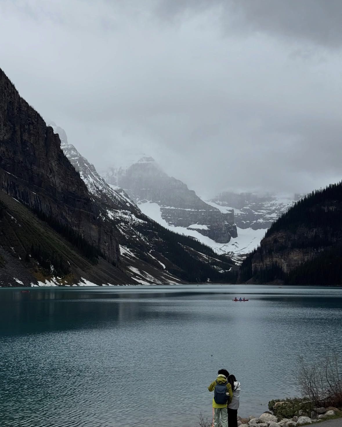 Two visitors at the water's edge below snowfields on an overcast day