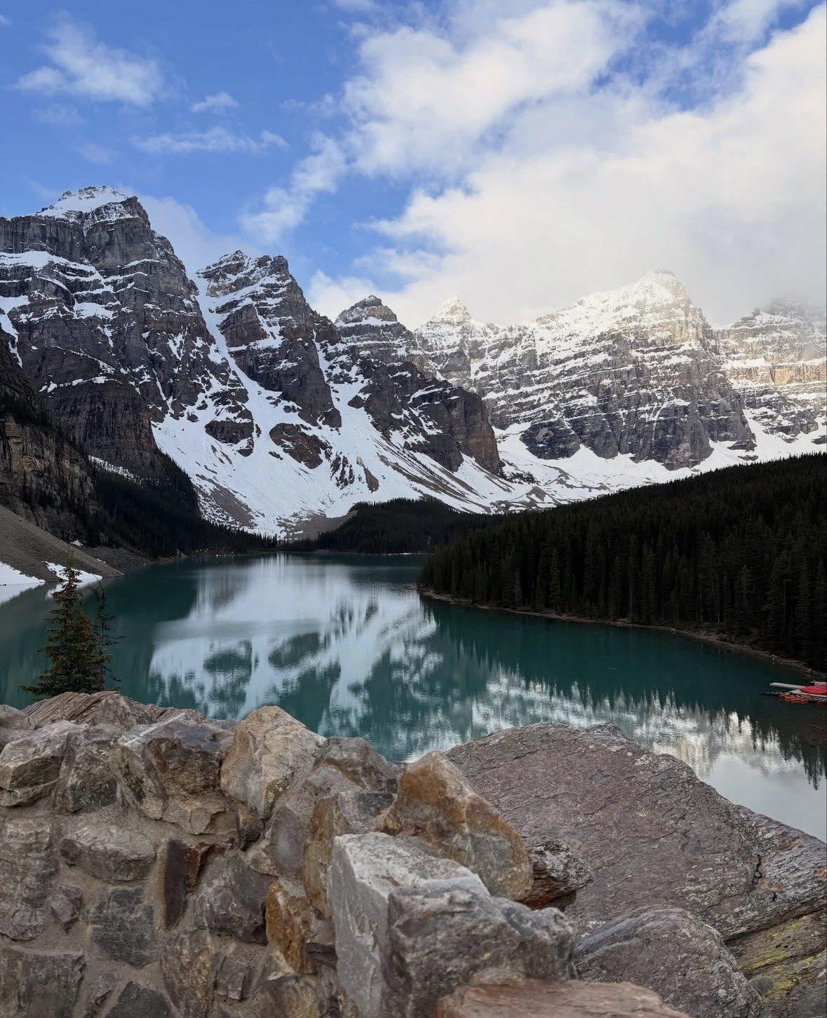 Snow-streaked peaks and forest mirrored in green water seen over a stone wall