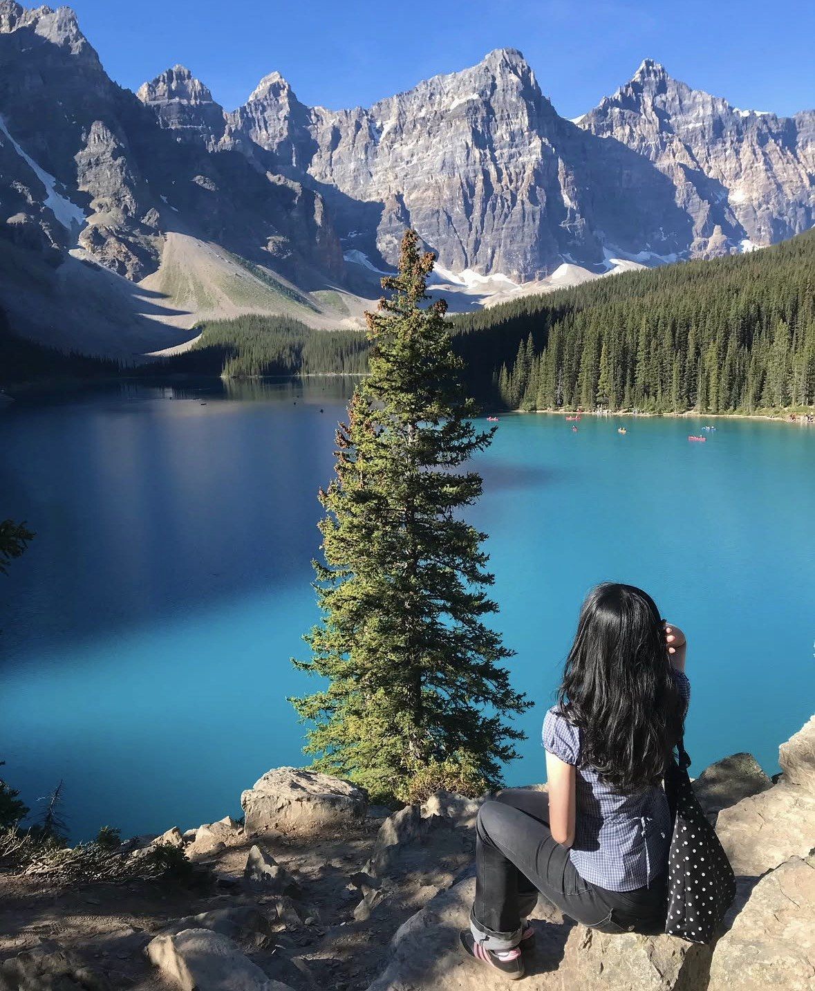 Woman seated on a rocky ledge above bright blue water and jagged peaks
